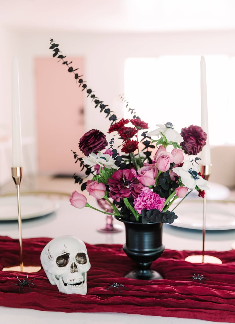 Elegant table setting with a floral arrangement featuring pink and burgundy flowers in a black vase, flanked by gold candlesticks and a white skull on deep red fabric.