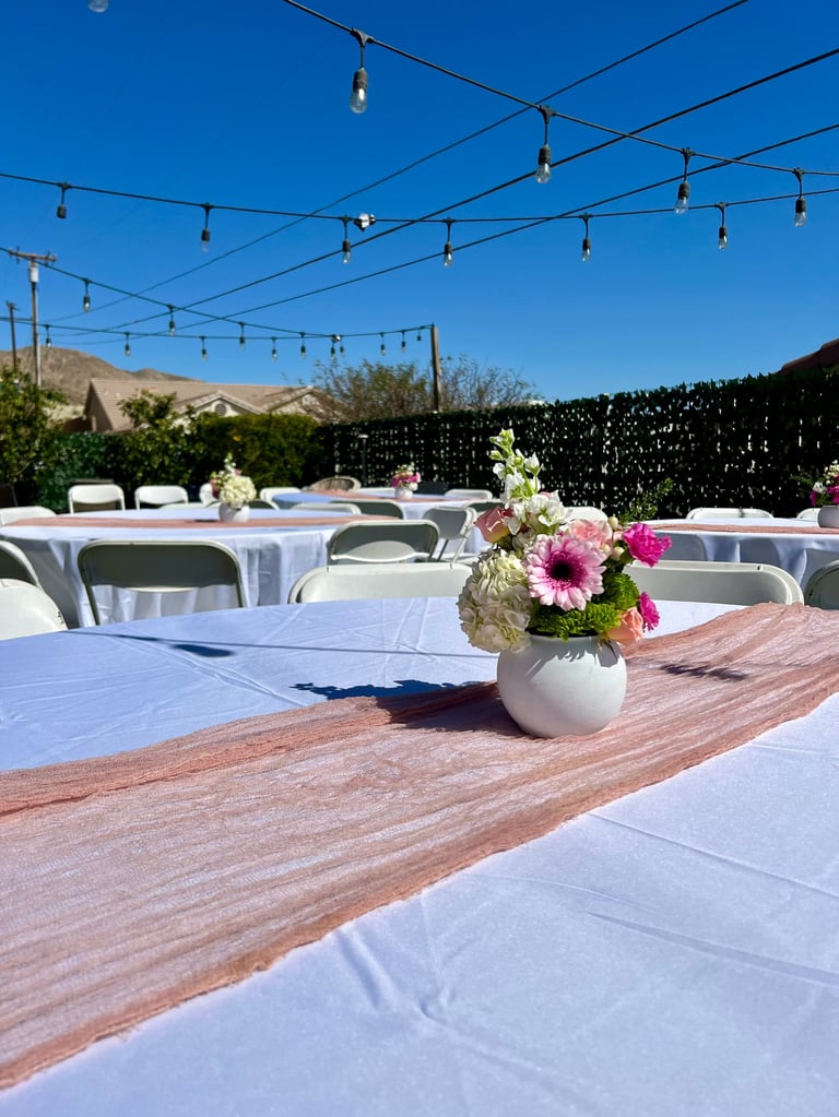 Outdoor event venue with white-draped tables, pink and magenta flowers in vases, string lights overhead, mountains and black privacy fence visible in background