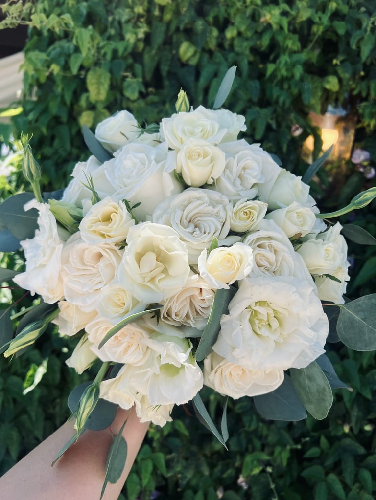Hand holding a bridal bouquet of cream and white roses with eucalyptus greenery against a leafy green background