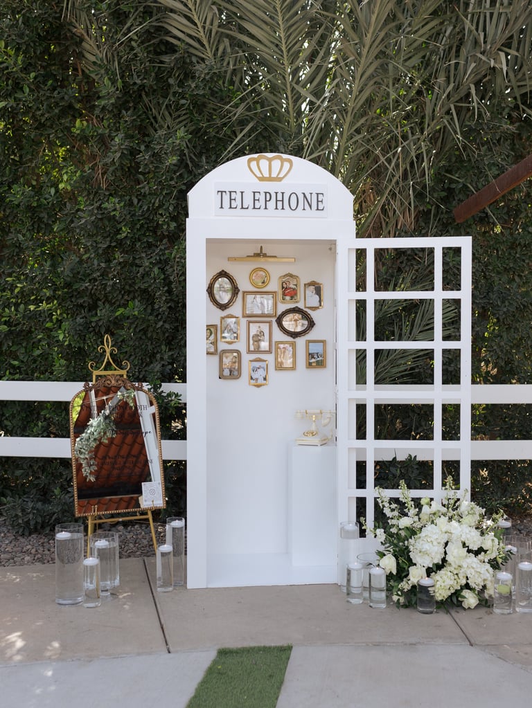 Vintage-style white telephone booth decorated with framed photos, a decorative lattice window panel, flowers, and festive garland arrangement