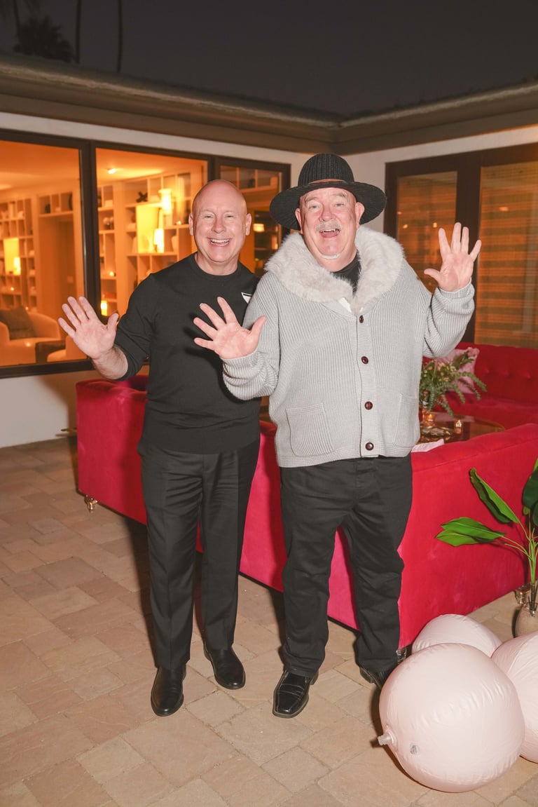 Two men waving at the camera in an indoor venue with red seating and warm lighting
