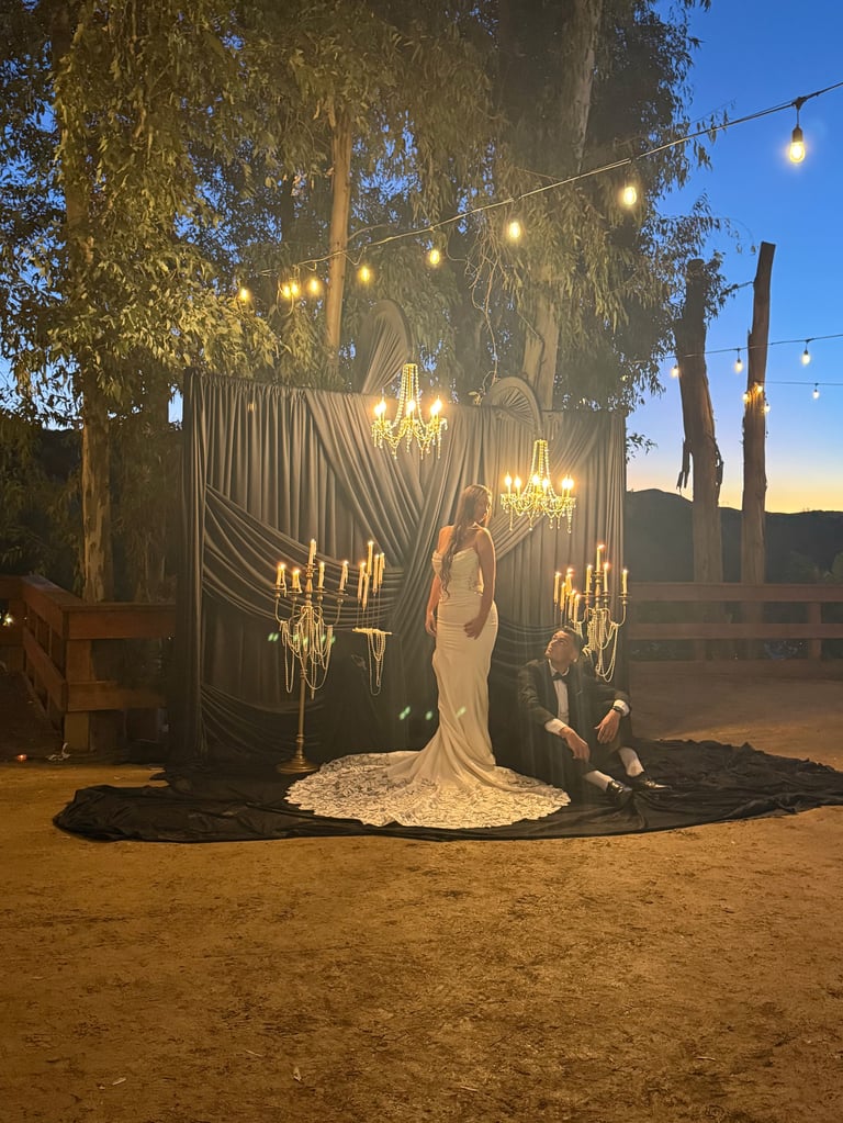 Outdoor wedding ceremony at dusk with bride in white dress, groom kneeling, elaborate backdrop with drapes and chandeliers under eucalyptus trees