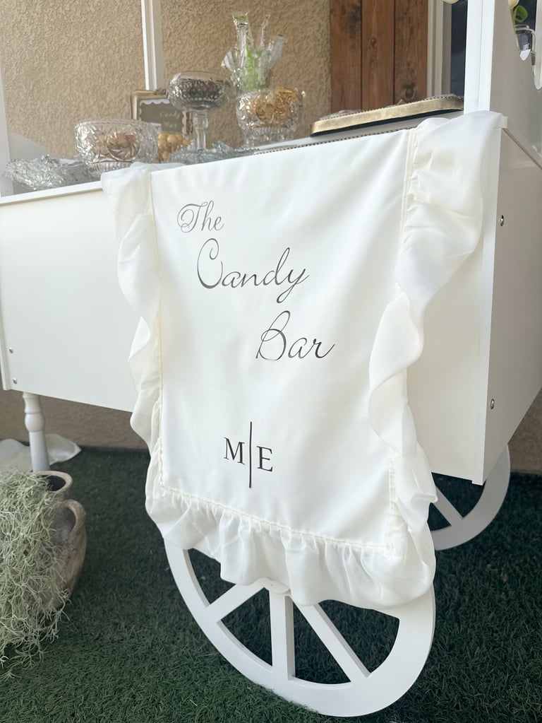 White cart draped with cream fabric labeled The Candy Bar decorated with glass jars of candy and decorative items