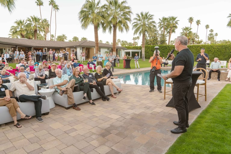 Outdoor event at a resort with guests seated around a pool area surrounded by palm trees, a man standing and speaking to the group