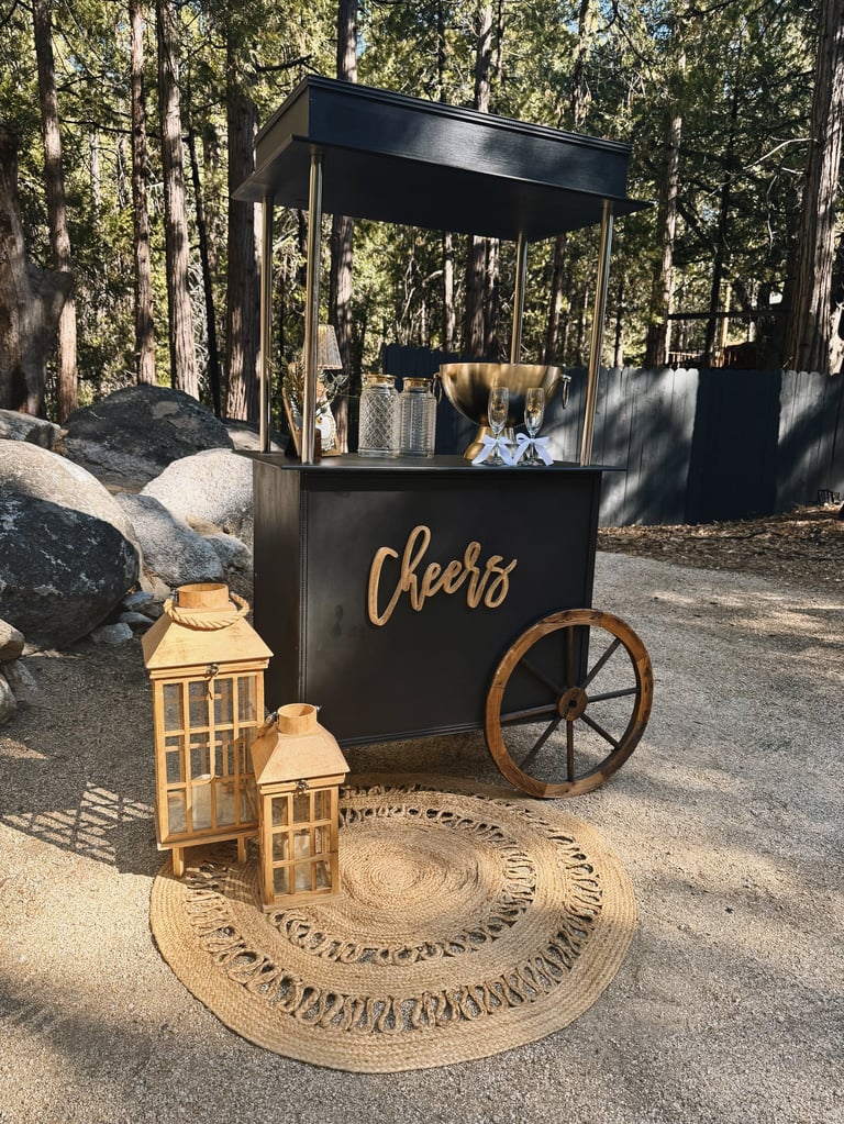 Black vintage-style cheers bar cart with wooden wheel in a forest setting, decorated with lanterns and woven rug