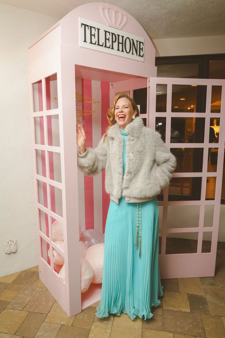 Woman in turquoise dress and grey fur coat posing in front of a pink and white British telephone box booth