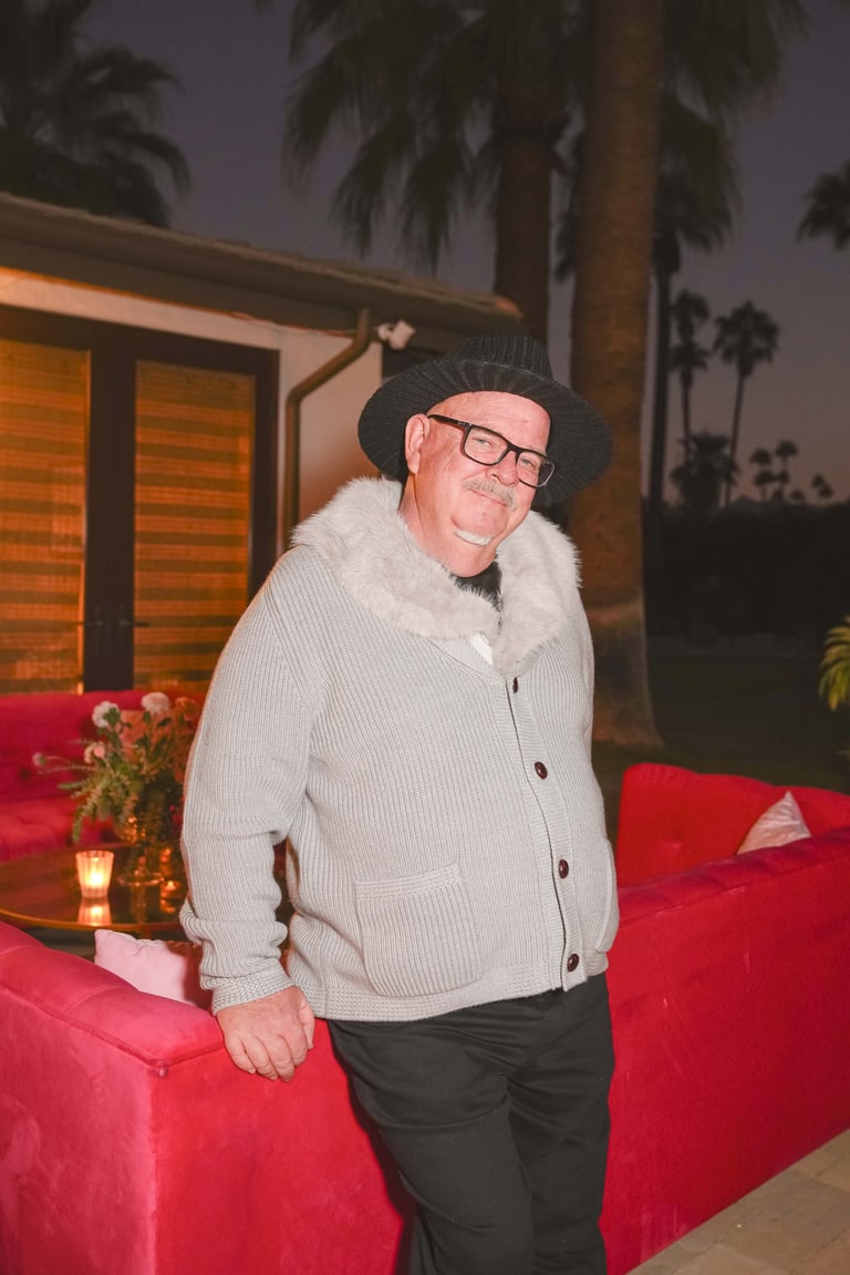 Man wearing glasses and a dark beanie posing in front of red furniture with palm trees visible in the background at dusk