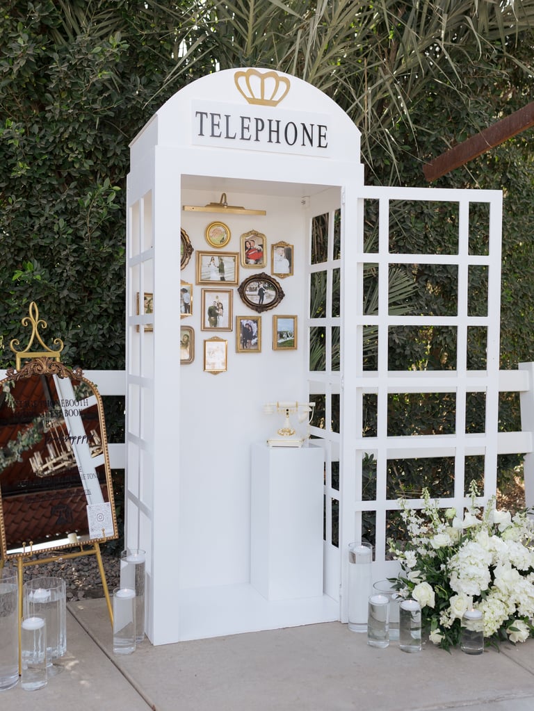 White vintage-style telephone booth decorated with framed artwork and gold crown emblem, surrounded by white flowers and seating area