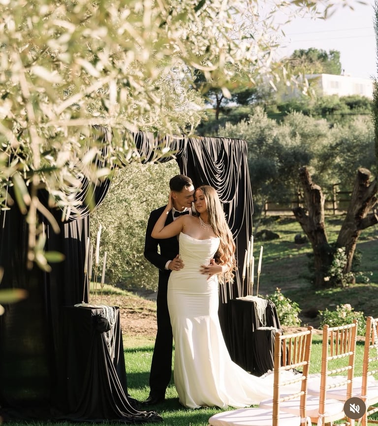 Bride and groom kiss under black and white striped wedding arch in olive grove with white flowers