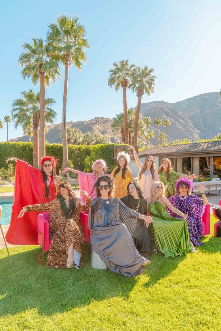 Group of women in colorful outfits posing on lawn with giant pink chair, palm trees, and mountains in background