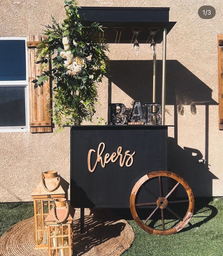 Black mobile bar cart with Cheers script lettering, wooden wheel, climbing greenery