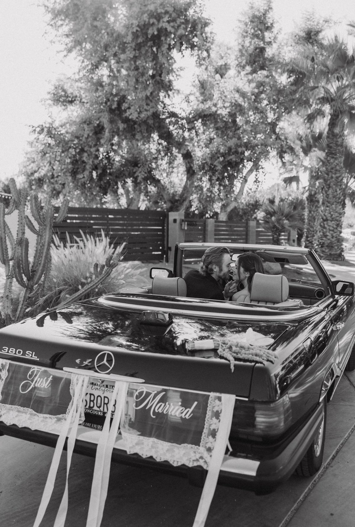 Black and white photo of a convertible Mercedes with Just Married ribbons, couple driving through palm tree-lined street