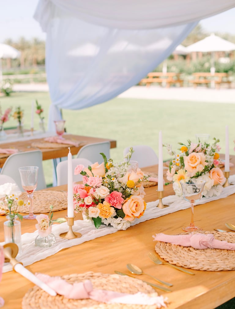 Outdoor wedding reception table with peach and pink floral centerpieces, white candles, place settings, and sheer blue draping overhead