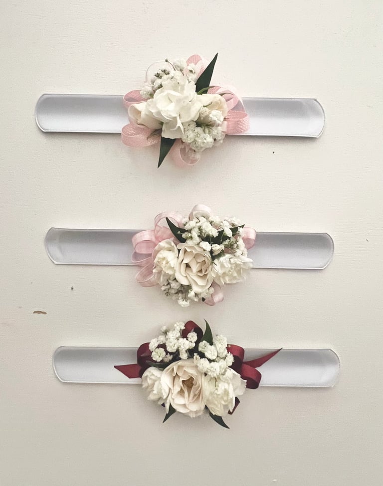 Three decorative wrist corsages with silver bands and floral arrangements in white, pink, and burgundy tones displayed vertically