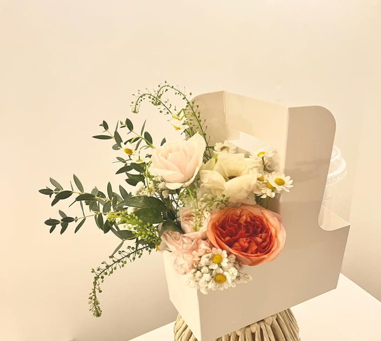 Elegant floral bouquet with cream roses, red garden rose, daisies, and green foliage in a cream-colored wrapper against a beige background.