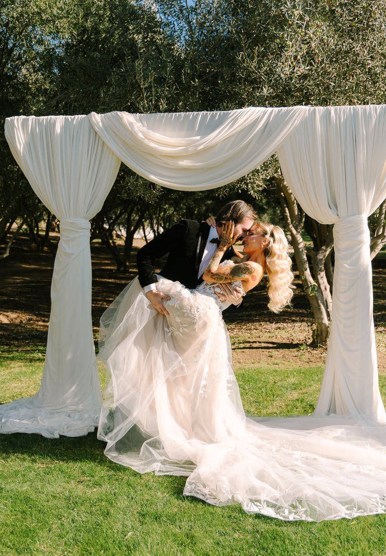 Groom dips and kisses bride under white draped ceremony arch in park with trees in background