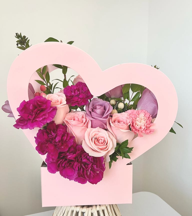 Pink heart-shaped floral arrangement with magenta carnations, roses, and greenery in a vase