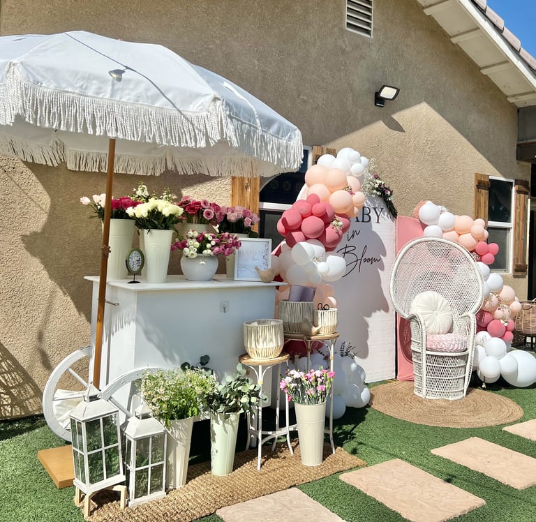 Outdoor event setup with white fringed umbrella, pink and white balloon arch, white shelving displaying flowers, plants, and decorative items on decorative patio