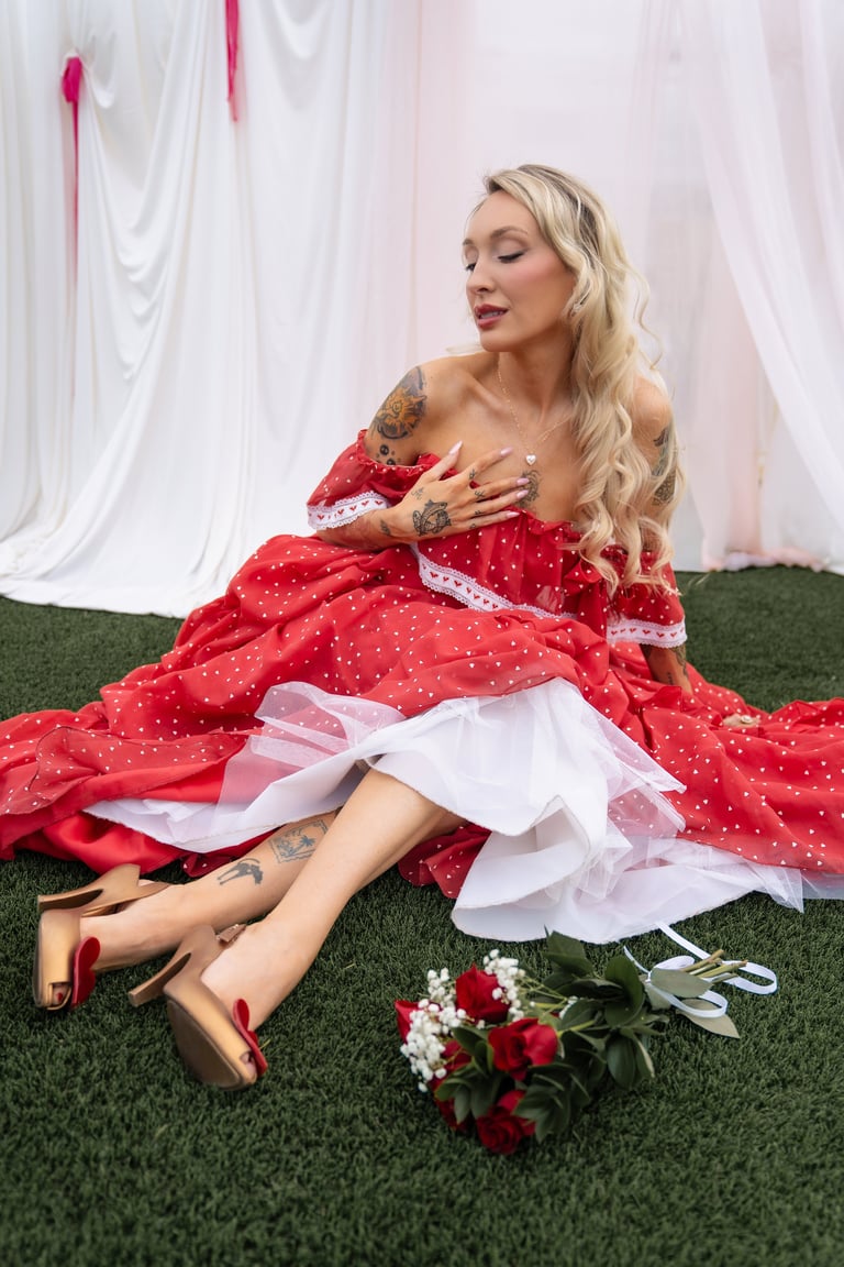 Woman in red off-shoulder gown sitting on grass with bouquet of red and white flowers against white backdrop