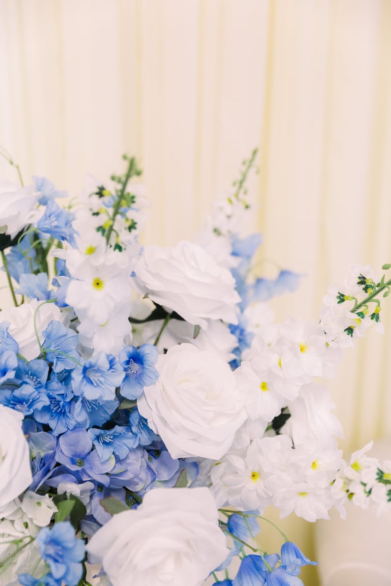 Elegant floral arrangement with white roses, blue and white hydrangeas, and yellow-centered daisies against a beige curtain backdrop
