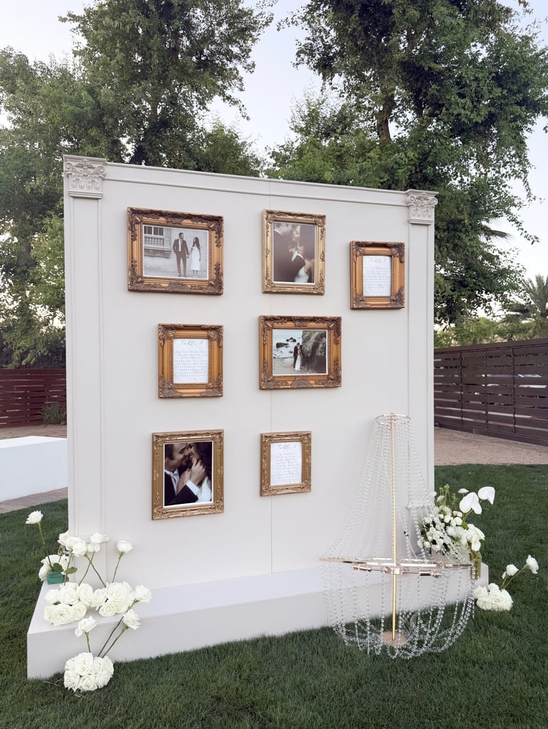 White backdrop wall with decorative ornate gold frames displaying photos, surrounded by white flowers and hanging paper chains in a garden setting