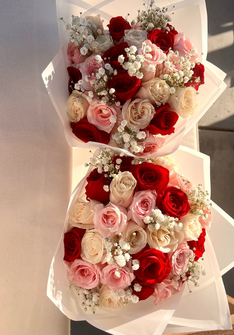 Two stacked bouquets of roses in red, pink, and cream with white babys breath accents, wrapped in white paper