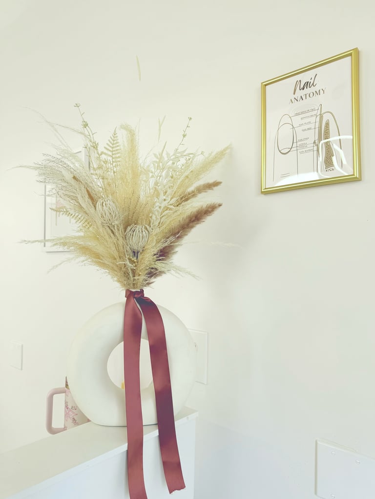 White vase with dried pampas grass and burgundy ribbon on shelf, with botanical wall art above