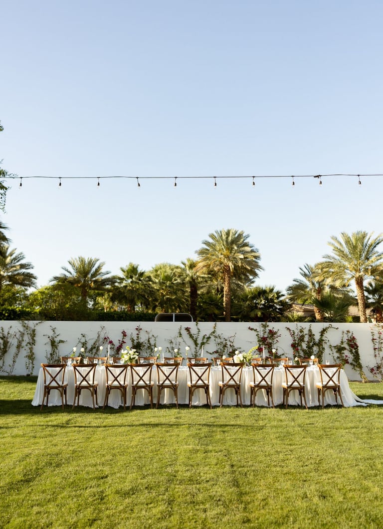 Outdoor wedding reception setup with decorated table, wooden chairs with fabric covers, palm trees, and string lights under clear sky