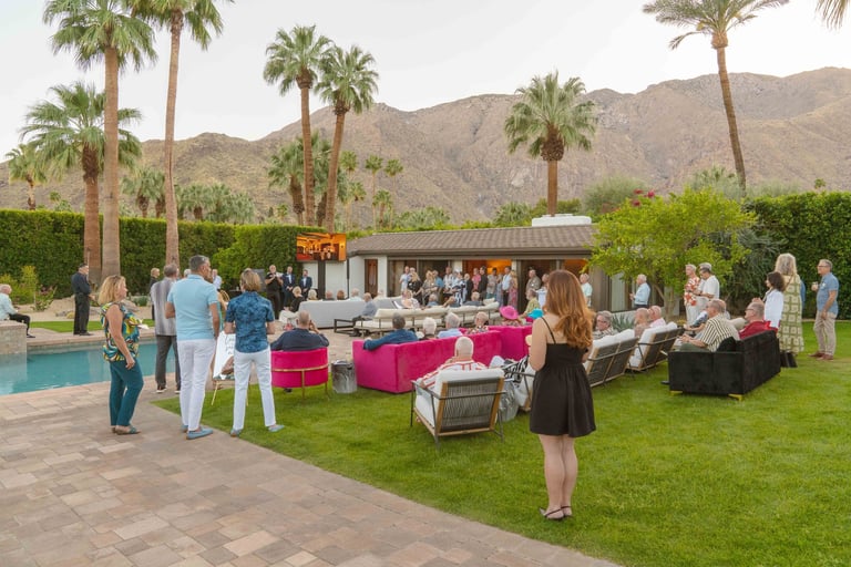 Outdoor garden party with guests socializing around pink furniture, pool, and palm trees with desert mountains in the background