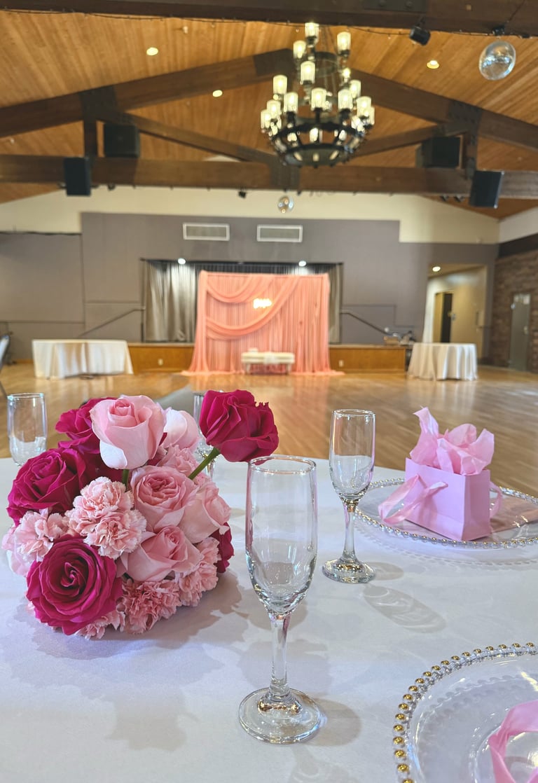Elegant banquet hall with pink and magenta floral centerpiece, place settings, and ornate chandelier above wooden-beamed ceiling