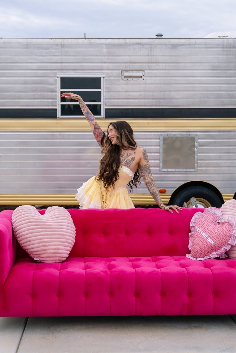 Woman with tattoos posing on a bright pink velvet couch in front of a silver and gold striped RV trailer