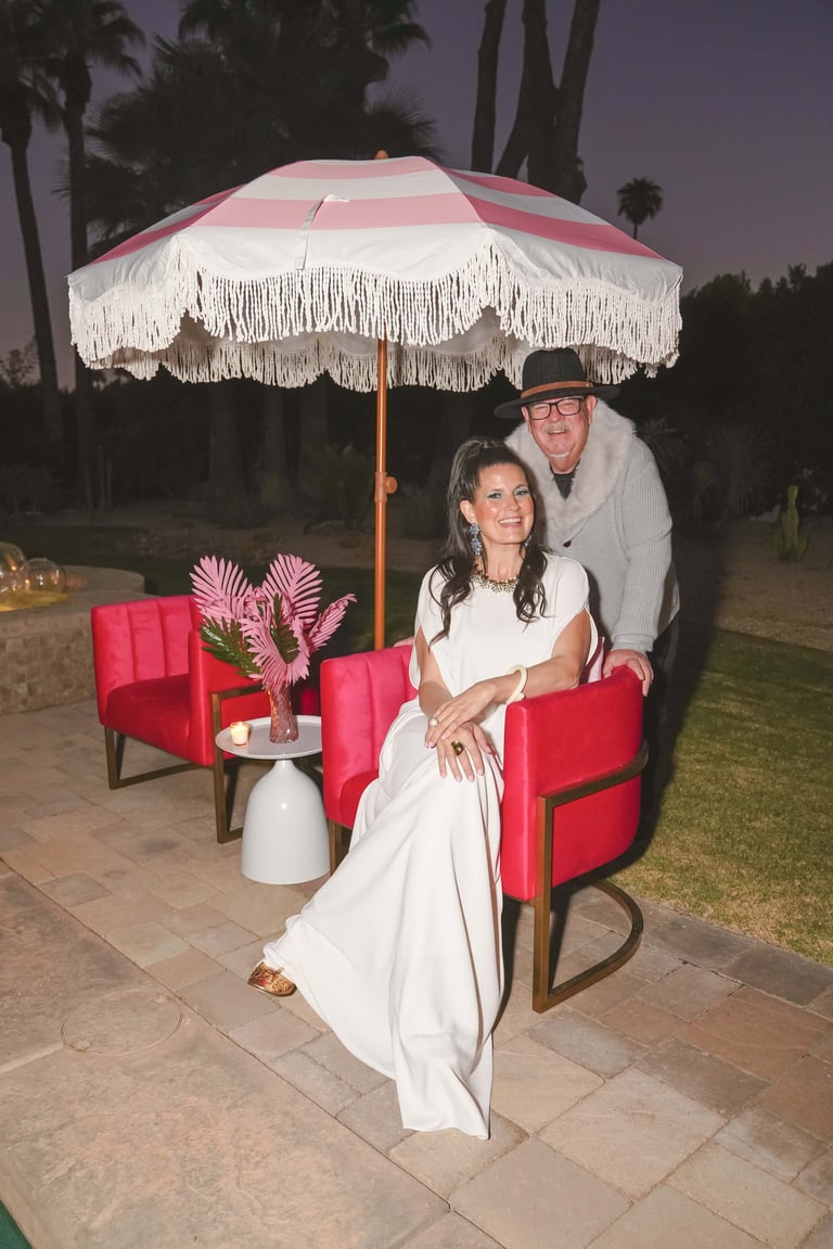 Couple posing under a pink and white striped umbrella with fringe on a patio at dusk, with red chairs and palm trees in background