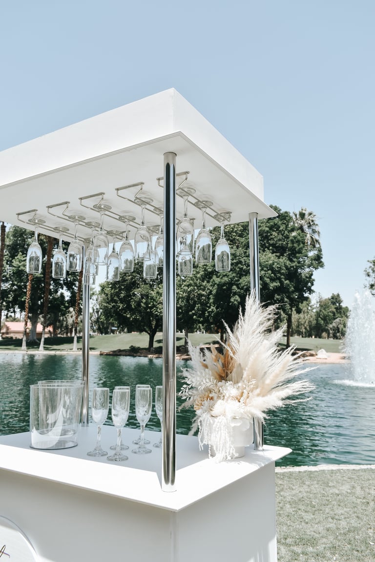 Modern white outdoor bar setup with hanging glassware, decorative pampas grass, and lake view in background