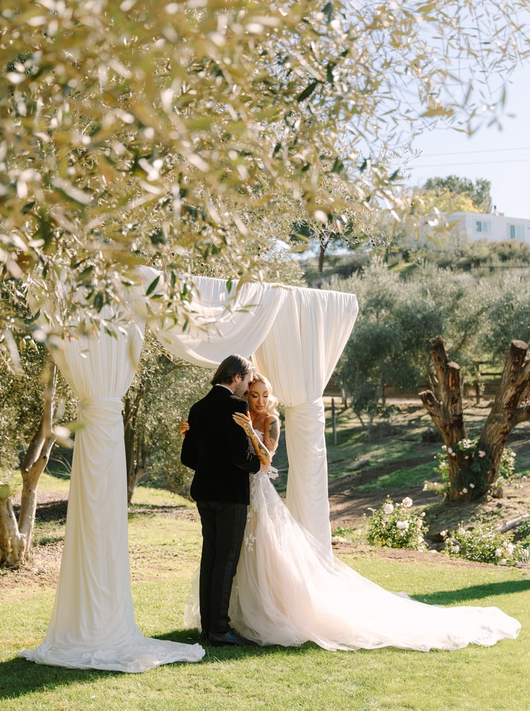 Bride and groom kissing under white draped wedding arch in olive grove setting