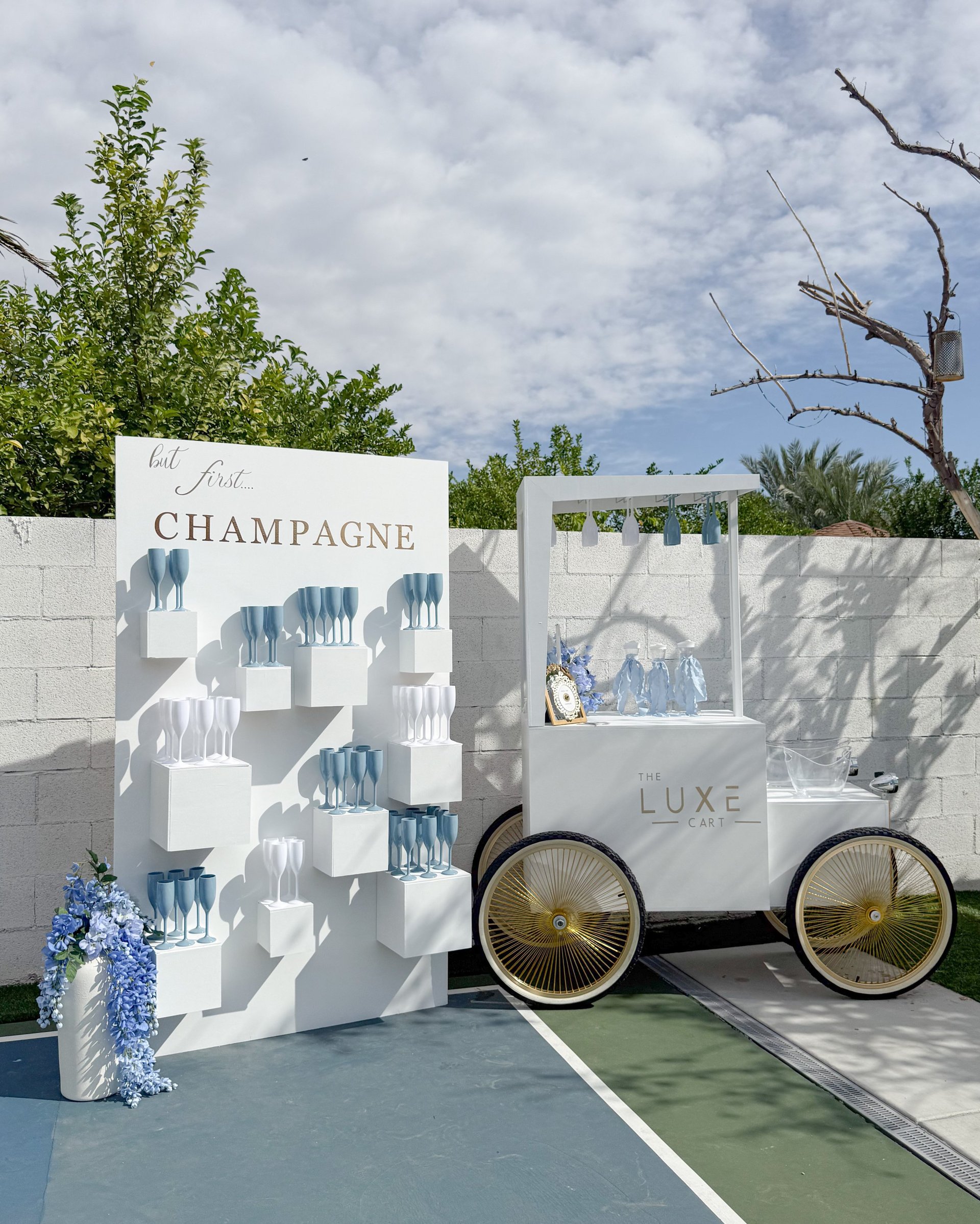 White luxury champagne cart display installation with decorative signage and blue flowers, set against white brick backdrop and garden greenery.