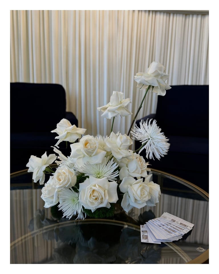 White floral arrangement with roses and gerbera daisies on a glass table in a modern office setting with black chairs and curtains in background