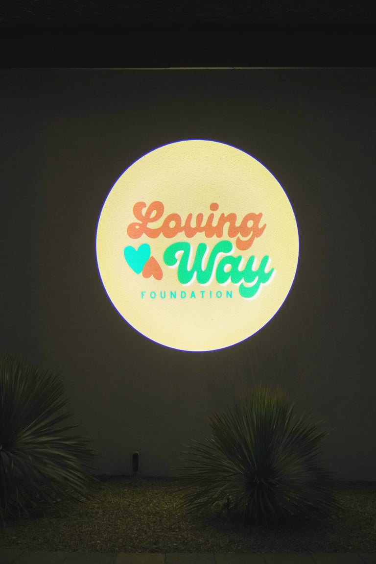 Neon circular sign displaying Loving Way Foundation logo with coral and turquoise text and heart icon on dark background with palm plants below