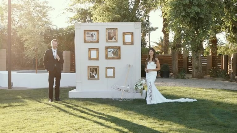 Bride and groom posing beside a white wall with framed photos in a garden wedding setting