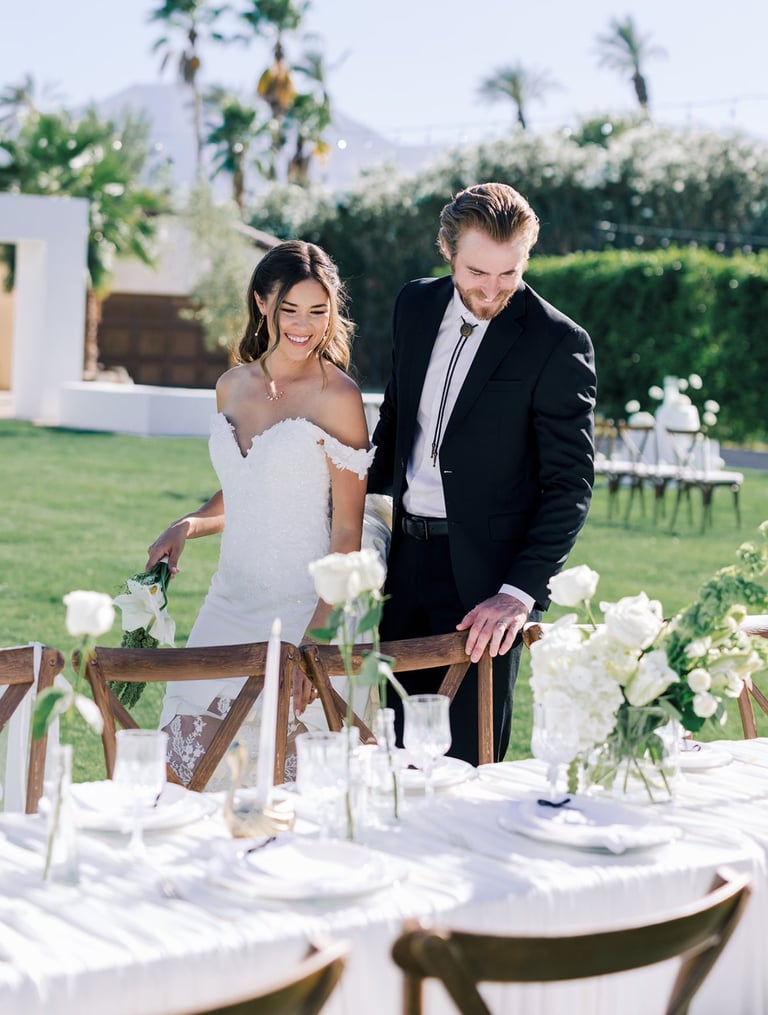 Bride and groom walking together at outdoor wedding reception table decorated with white flowers and place settings
