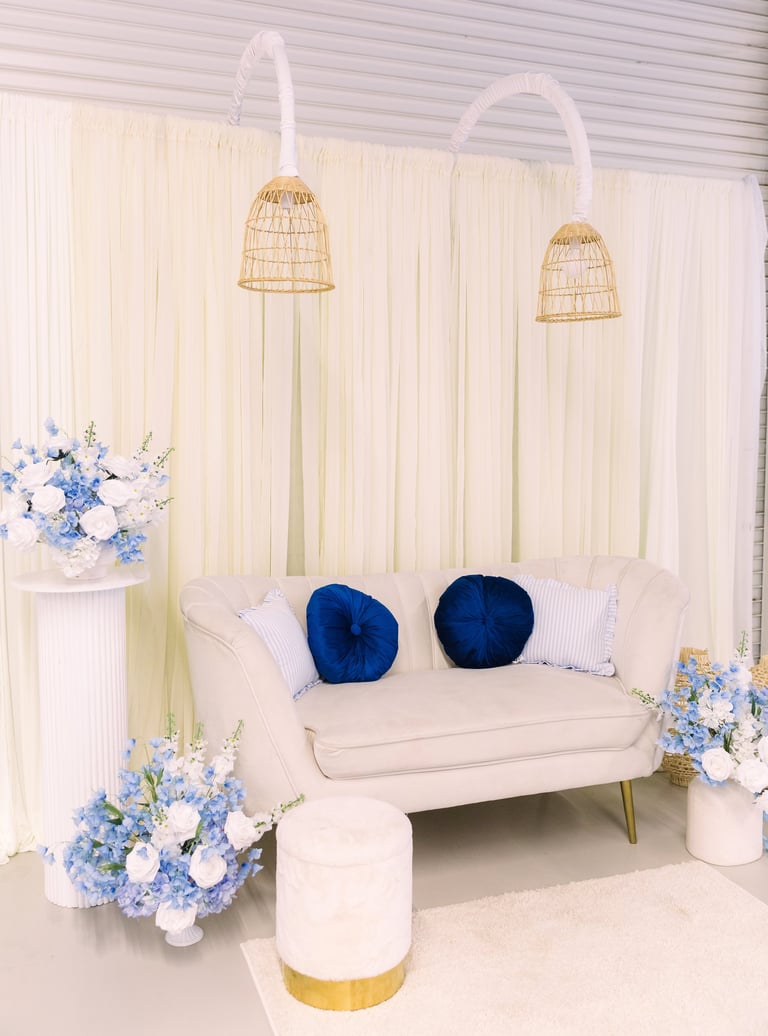 Elegant seating area with cream sofa, blue velvet pillows, woven pendant lamps, blue and white floral arrangements, and gold-accented ottoman