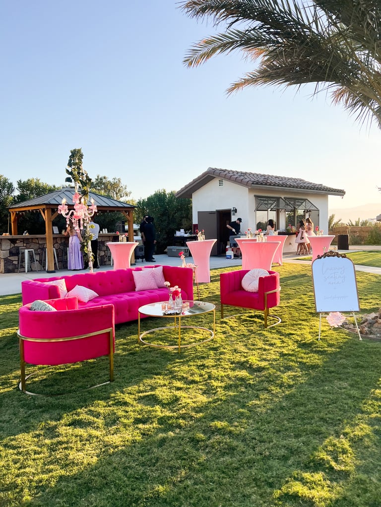 Outdoor event setup with bright pink furniture and cocktail tables on a green lawn, with palm trees