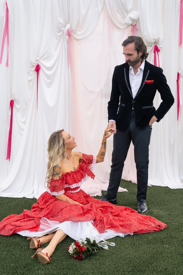 Man in black suit helping woman in red off-shoulder dress stand up on green grass with floral backdrop