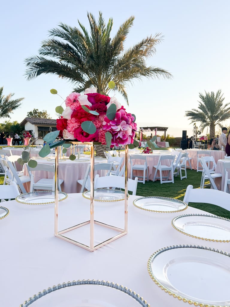 Outdoor wedding reception setup with pink and red floral centerpiece on geometric stand, elegant table settings, and palm trees in background