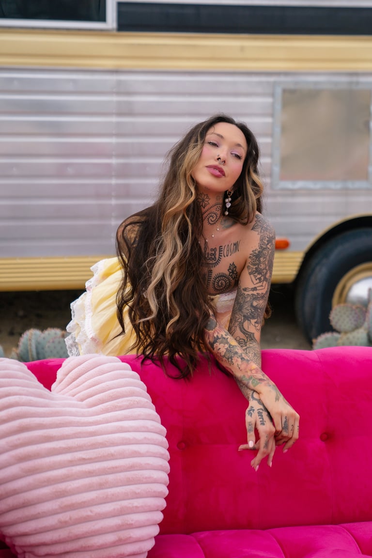 Tattooed woman with long dark wavy hair sitting on pink velvet couch in front of vintage trailer