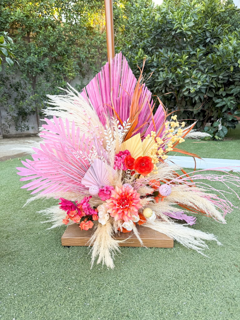 Colorful floral arrangement with pink palm leaves, red and coral flowers, dried grasses on wooden base in garden setting