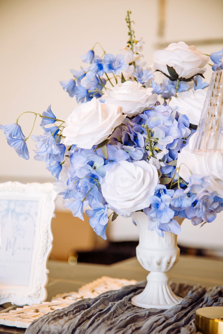 White and blue floral arrangement in a pedestal vase with white roses, delphiniums, and greenery