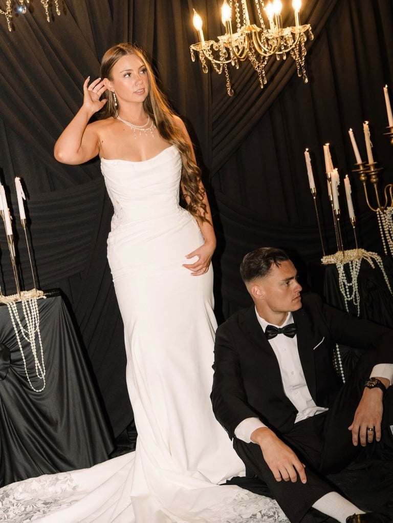 Woman in white wedding dress standing beside seated man in black tuxedo in elegant venue with chandelier and candles