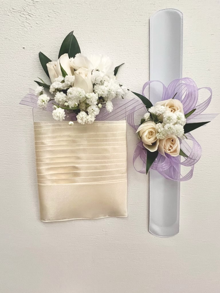 Two floral arrangements with white flowers, roses, and babys breath - one in a cream ribbon-wrapped container and one attached to a white pillar candle with purple tulle bow.