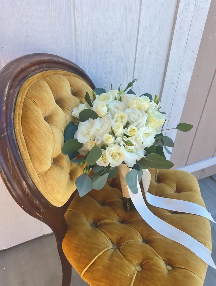 White and cream flowers with greenery tied with white ribbon resting on a golden yellow upholstered chair against a white wooden wall