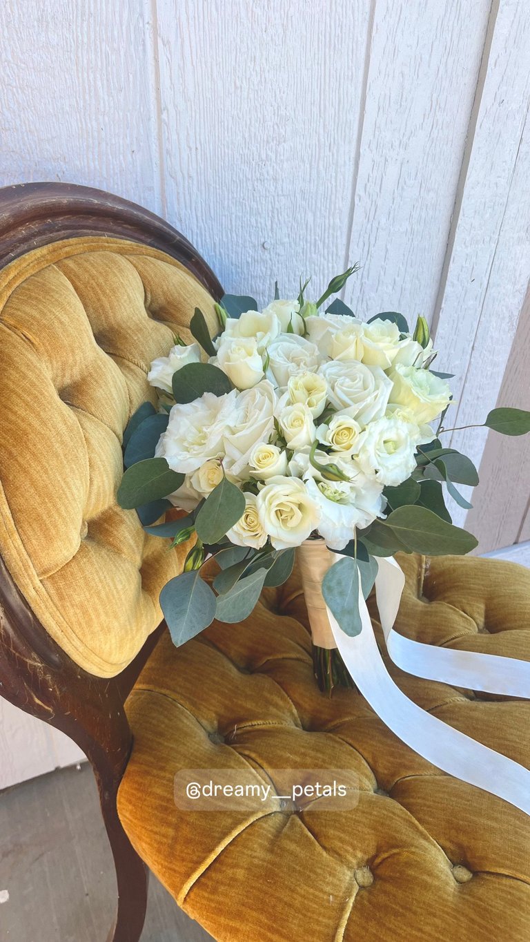 Bridal bouquet of white and cream roses with eucalyptus leaves on a gold velvet chair against white shiplap background